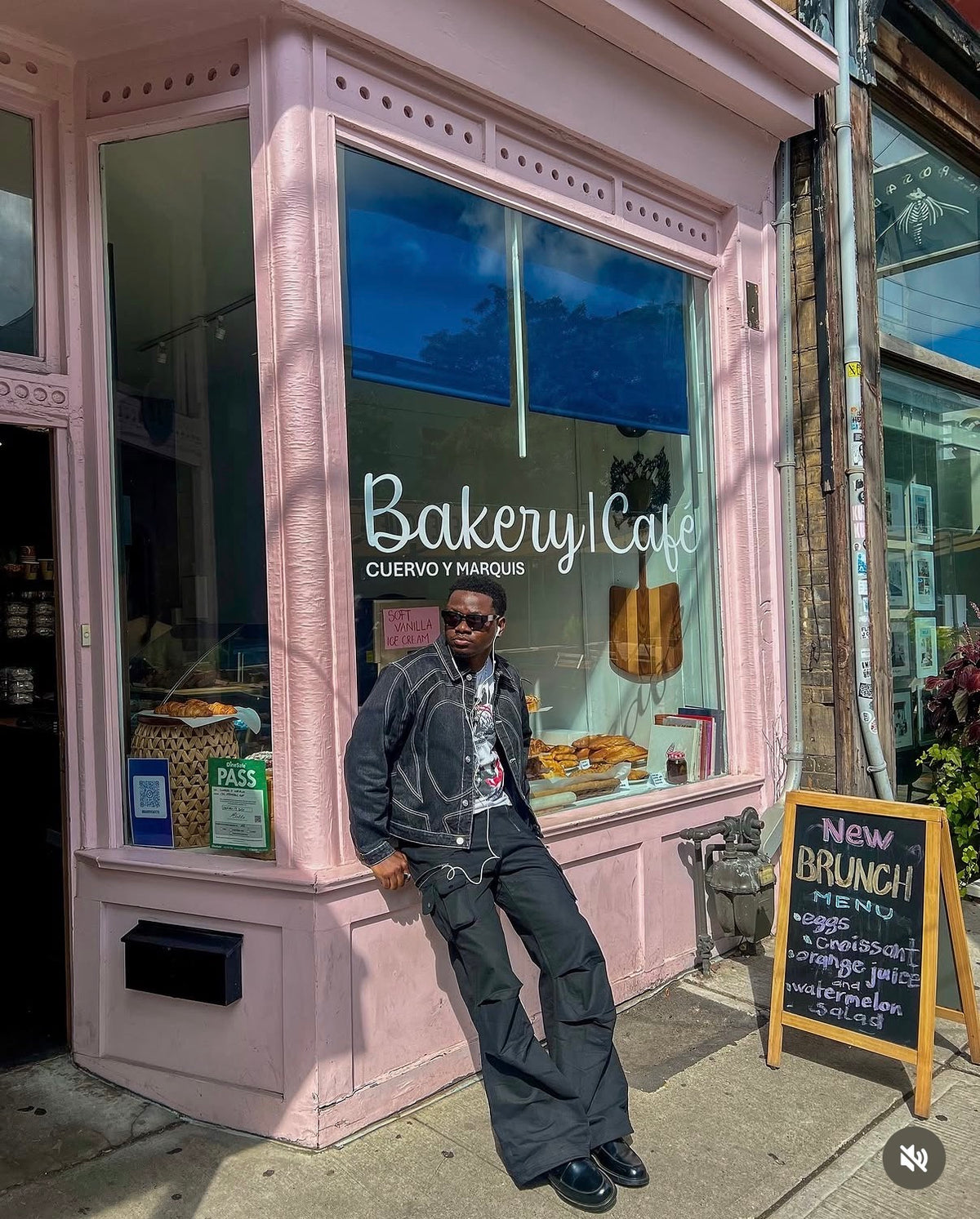Person wearing the black Midnight Voyager denim jacket styled with dark trousers outside a pink New York bakery storefront.