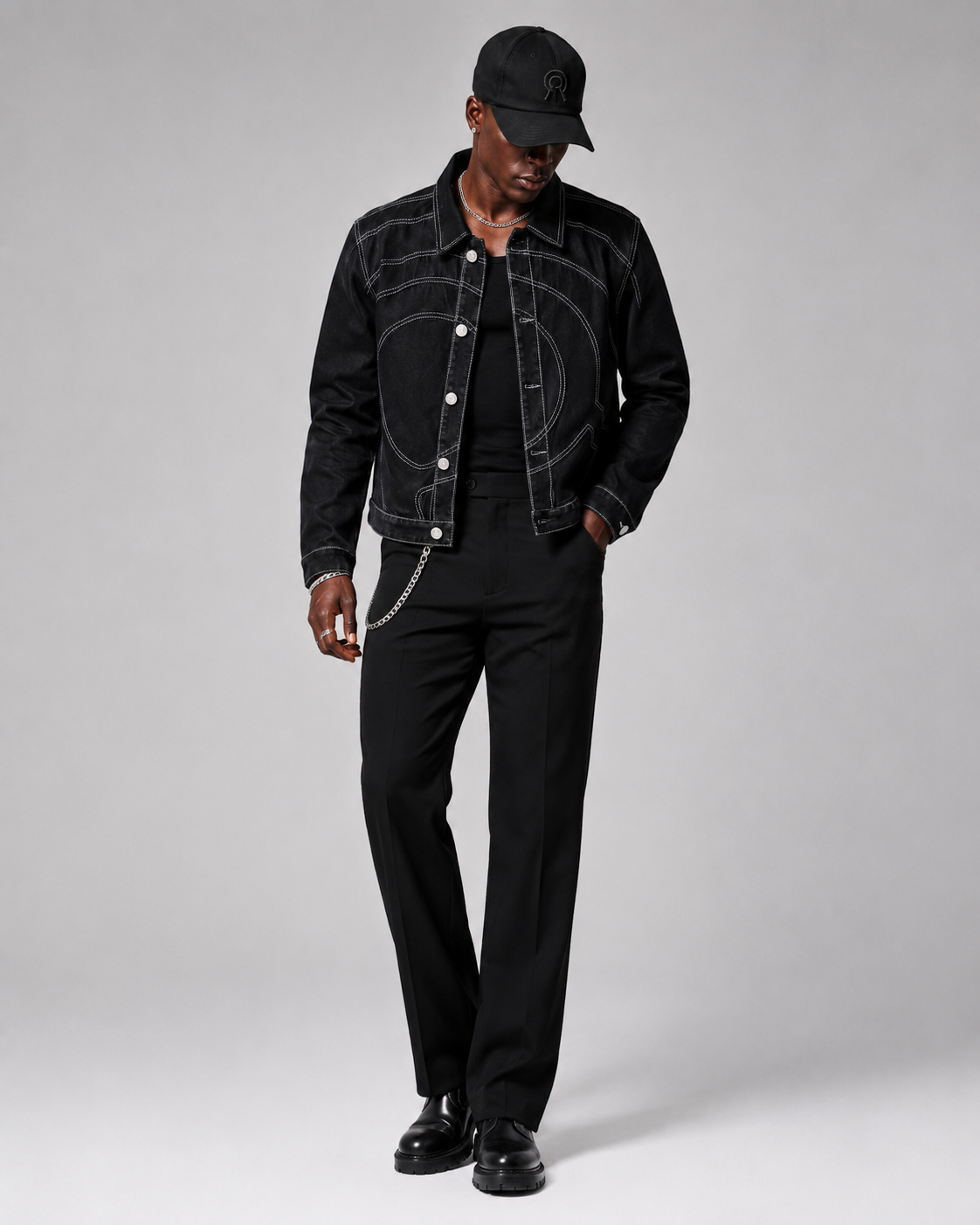 A model wearing a black denim jacket with white contrast stitching and a circular embroidered design on the chest, paired with black trousers, a black cap, and black shoes.