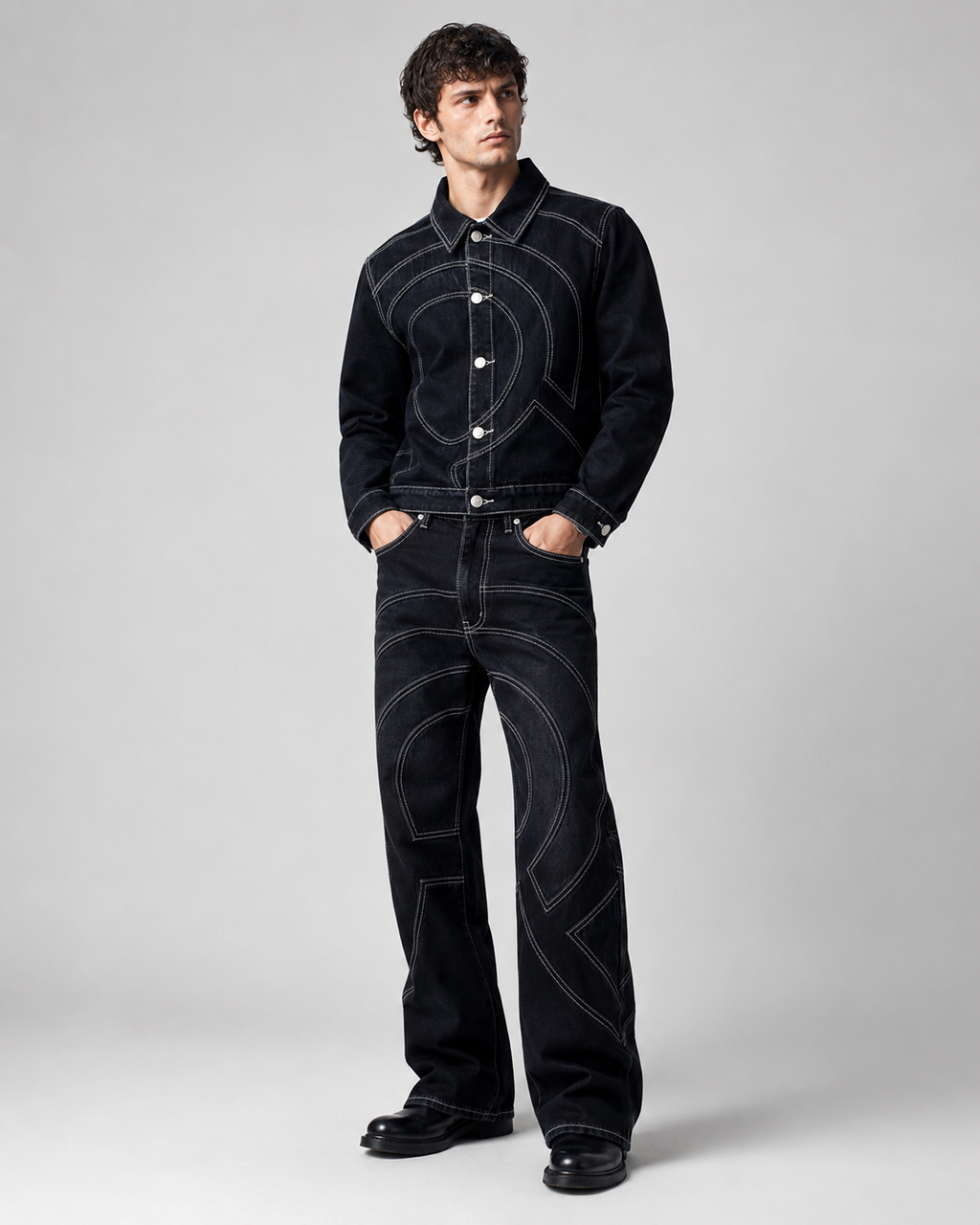 A man modeling a black denim jacket and matching flared leg jeans with prominent white contrast stitching.