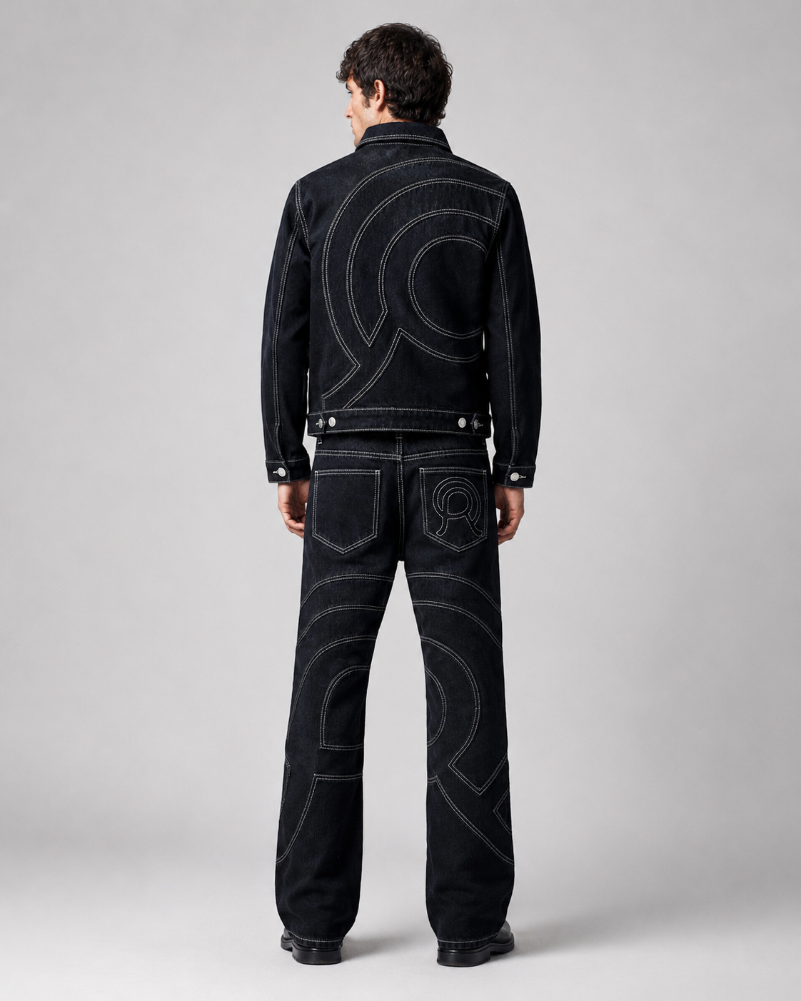 A model viewed from the back wearing a black denim jacket and matching flared leg jeans with large white contrast stitching forming a circular graphic pattern.