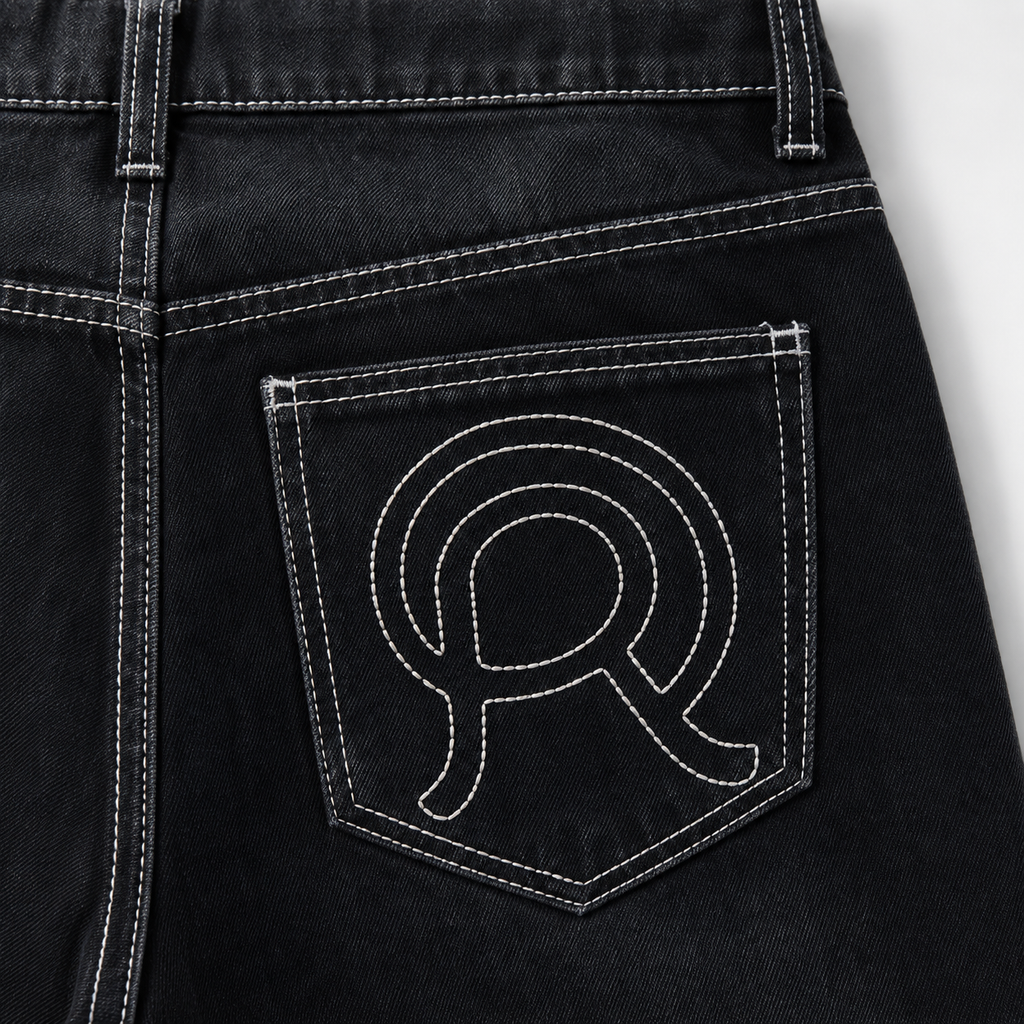Close-up of the back pocket of black denim jeans with white contrast stitching and a large embroidered white monogram logo on the pocket.